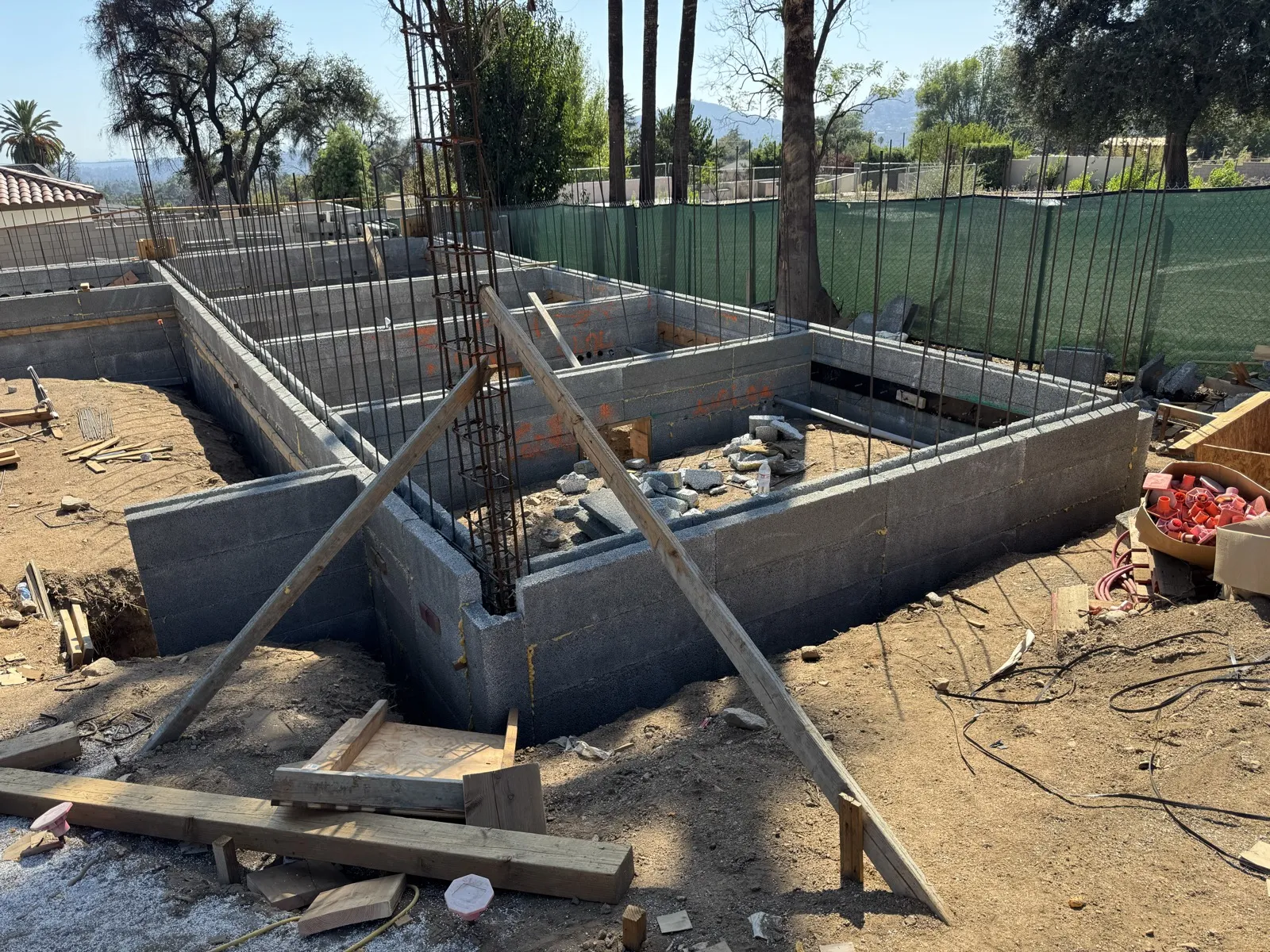 ICCF stem walls forming the raised floor house footprint with vertical rebar at the East Palm Street Altadena fire rebuild, wooden form bracing and green mesh fence with oaks and San Gabriel Mountains behind