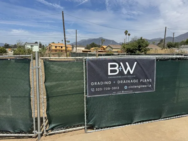Freshly rough graded Glenrose Avenue lot in Altadena during fire rebuild, with B+W Engineering grading and drainage plans sign on construction fencing and San Gabriel Mountains behind