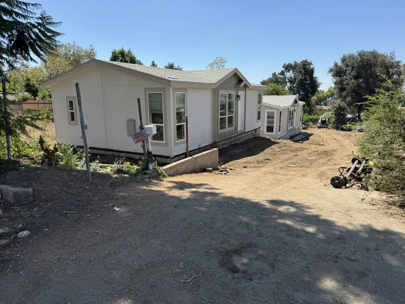 Newly placed white manufactured home on a graded dirt hillside lot in Altadena during an Eaton Fire rebuild, with a low concrete retaining wall, mailbox, Sigalert construction sign on a metal post, and a red mini skid steer parked at the edge of the yard