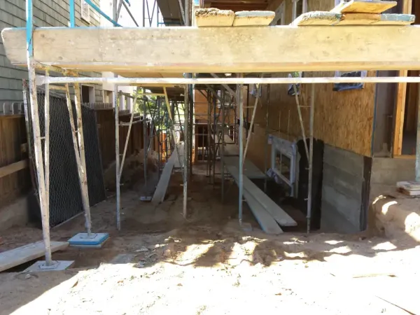 Metal scaffolding, OSB wall sheathing, and concrete foundation with waterproofed basement window during early-stage grading and excavation