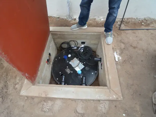 Below-grade concrete utility vault installation with open metal access door, black basin lid, plumbing connections, and electrical wiring for sump pump system