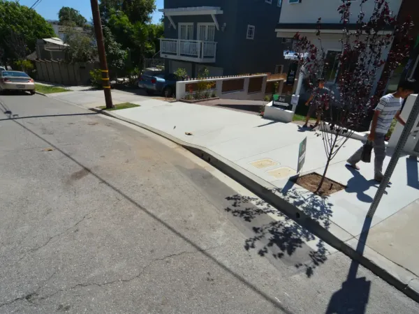 Newly constructed concrete sidewalk and driveway apron on steeply graded residential street with modern curb and gutter system, drainage scuppers, and tree well