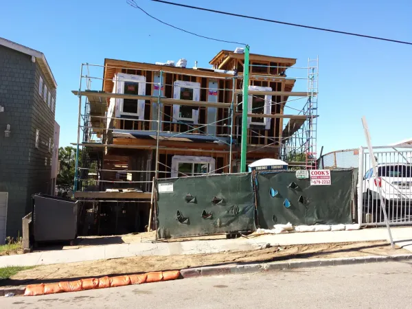 Residential construction site with multi-story wood-frame house, scaffolding, temporary green mesh fencing, and orange sediment control boom at street curb