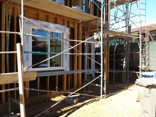 Wood framing and structural details of building under construction with installed white-framed window, MOISTOP flashing, metal scaffolding, and uneven dirt grading